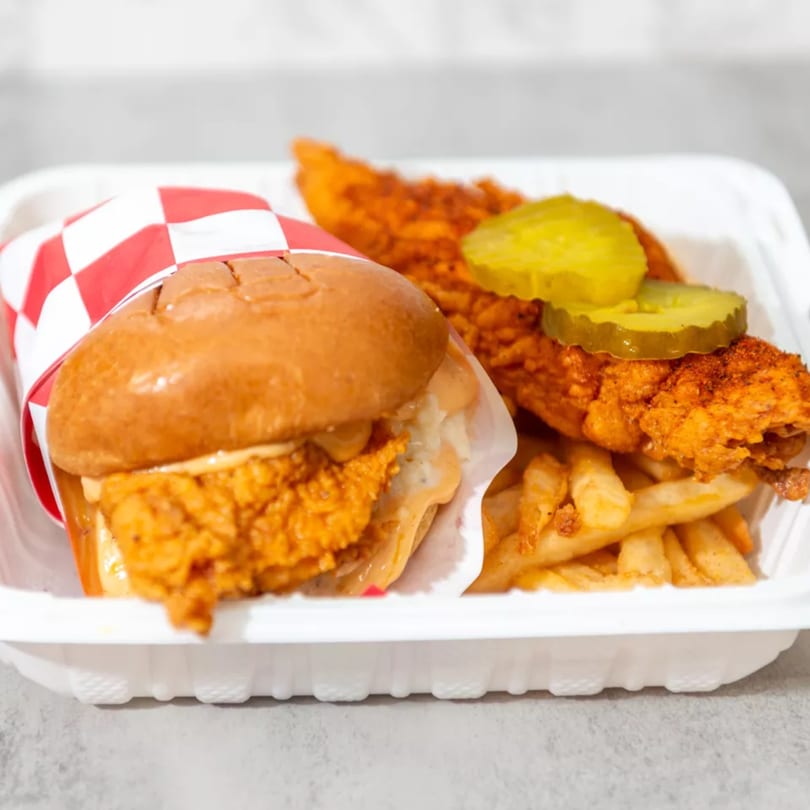Spicy fried chicken sandwich, chicken tenders with pickles, and french fries in a white takeout container.