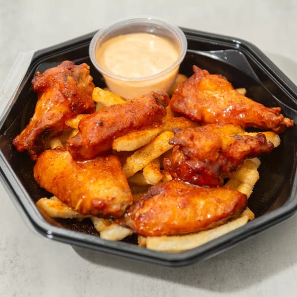 Spicy chicken wings and french fries in a black takeout container with a side of dipping sauce.